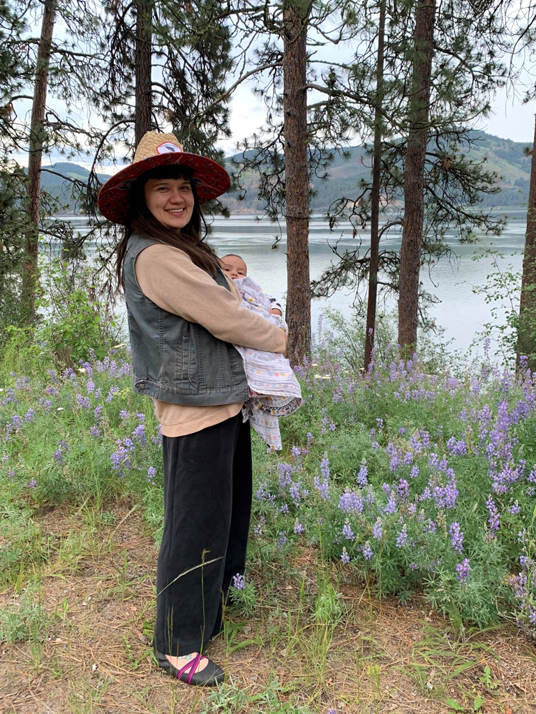 Emma standing beside a lake and patch of flowers dressed in black pants, tan sweatshirt, denim vest, and woven hat carrying her son wrapped in a blanket.