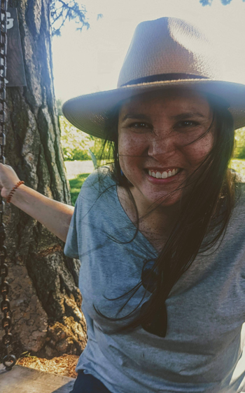 A woman wearing a brown hat and grey shirt smiling at the camera while on a wood and chain swing hanging from a tree. 