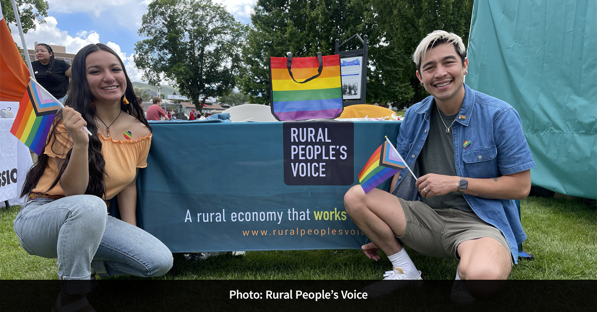Two individuals standing in front of a tabling booth for Rural People’s Voice, holding Pride flags.