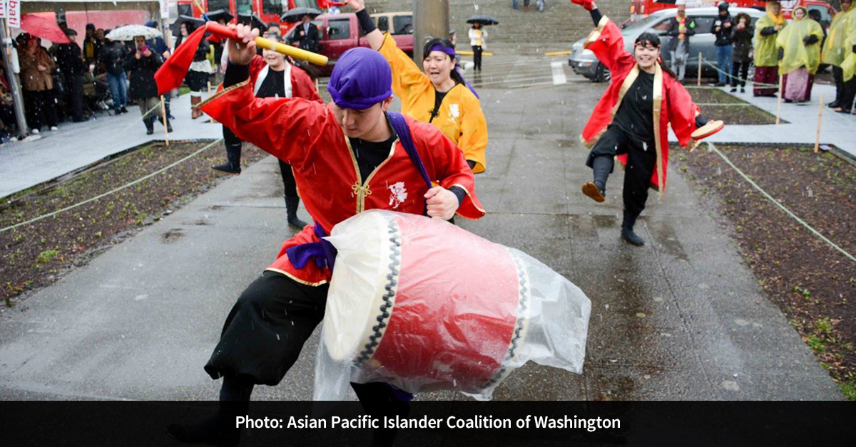 Four individuals wearing outfits of red, yellow, black, and purple, performing an outdoor ceremony with drums in the rain.