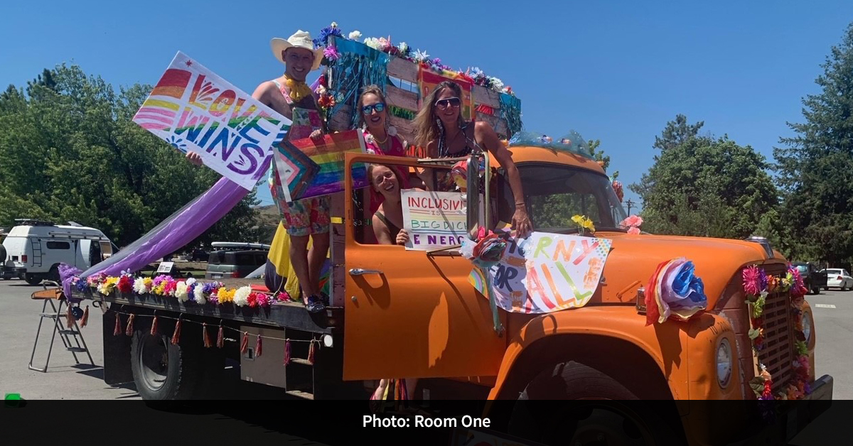 Four people holding Pride signs and flags in an orange truck decorated with colorful flowers.