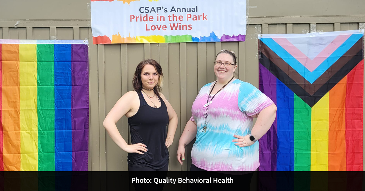 Two people standing beside Pride flags and a sign that reads, “CSAP’s Annual Pride in the Park Love Wins.”