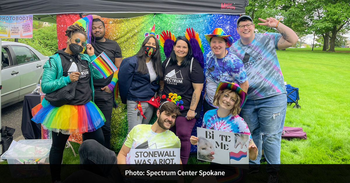 Eight people standing in front of a rainbow backdrop holding Pride signs and wearing rainbow accessories.