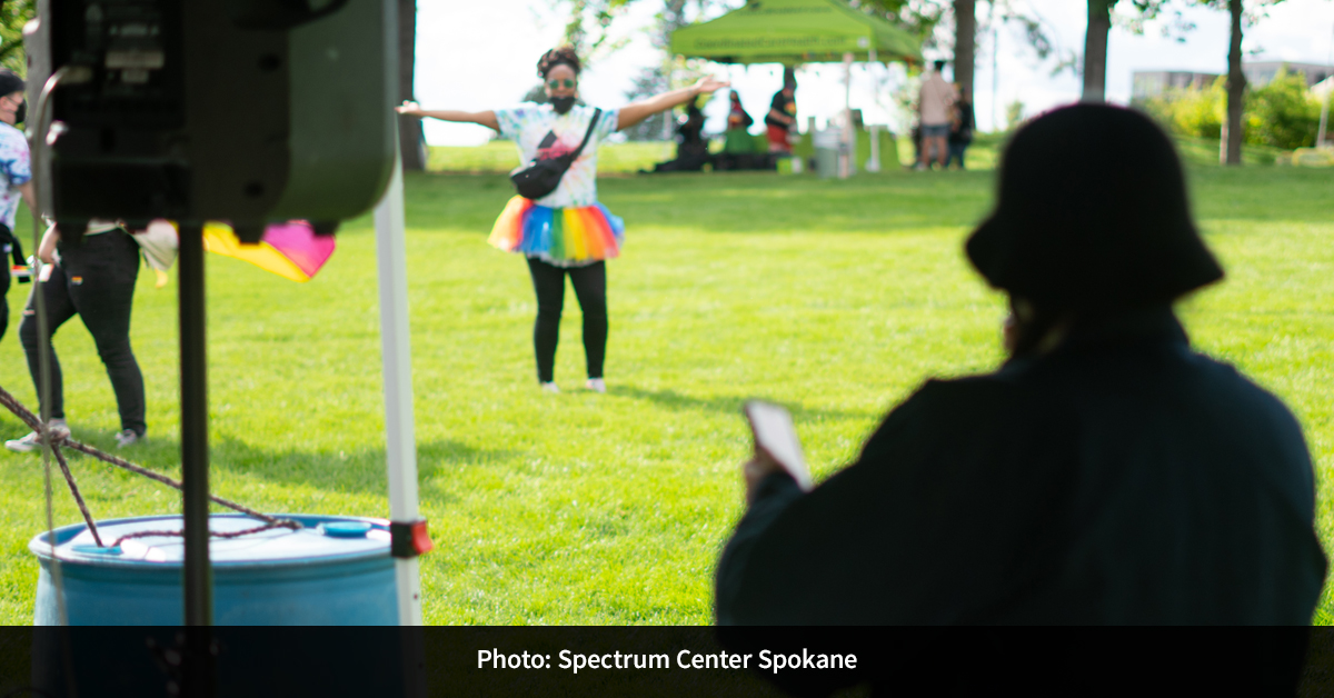 An individual in a rainbow tutu standing with their arms open in a grass field by near tabling booths.